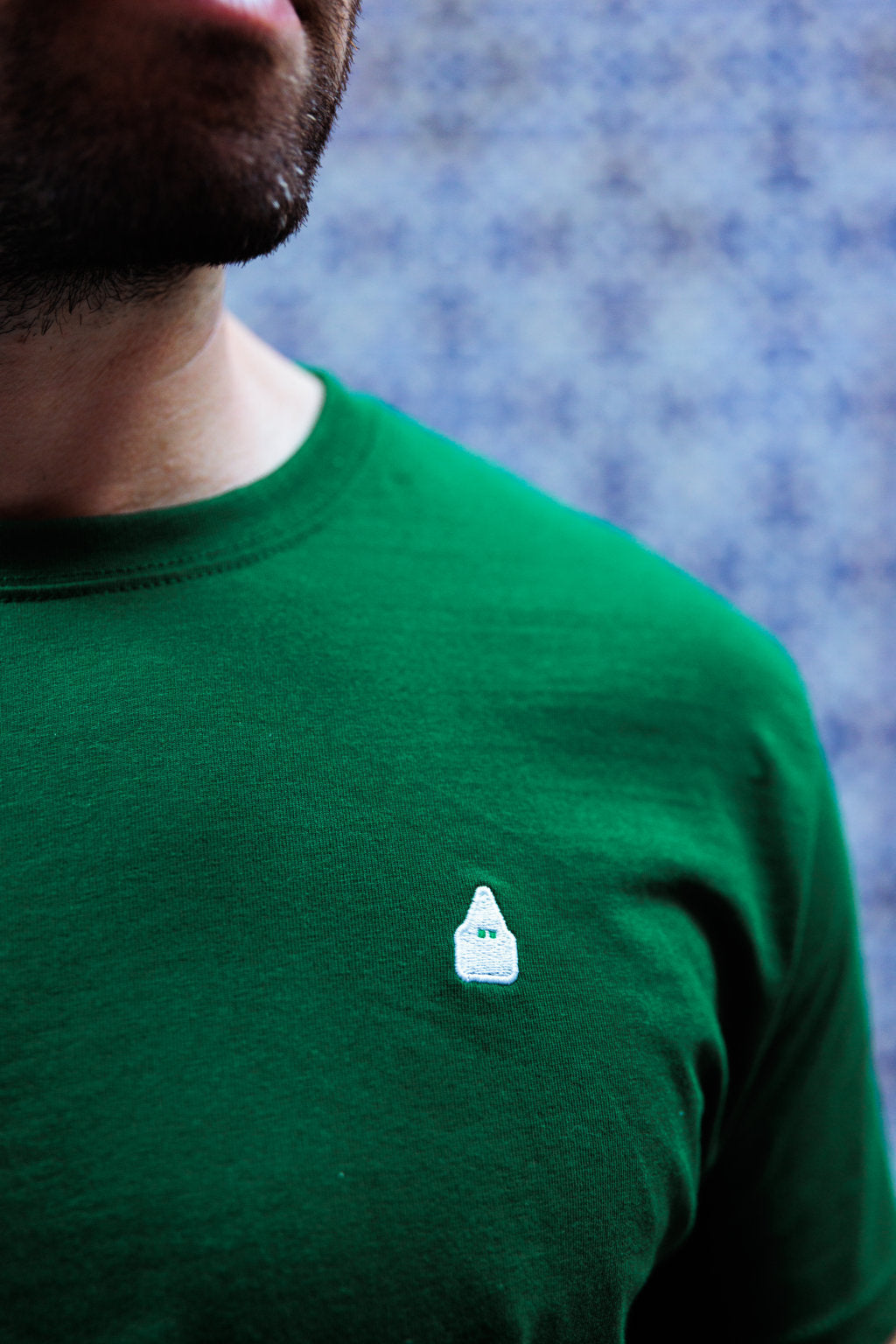 Primer plano del nazareno de TRAMO bordado en blanco sobre camiseta verde.