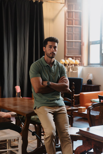 Hombre con polo verde de algodón TRAMO apoyado en una mesa y combinado con pantalón beige.