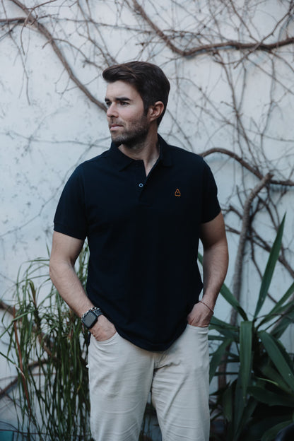 Hombre vistiendo polo azul marino TRAMO y pantalón beige en un entorno exterior con vegetación de fondo.